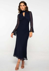 Vestido azul marino con mangas largas transparentes, cuello alto y un escote en forma de llave. Con un dobladillo acampanado y un material texturizado. Combinado con tacones dorados con correas.