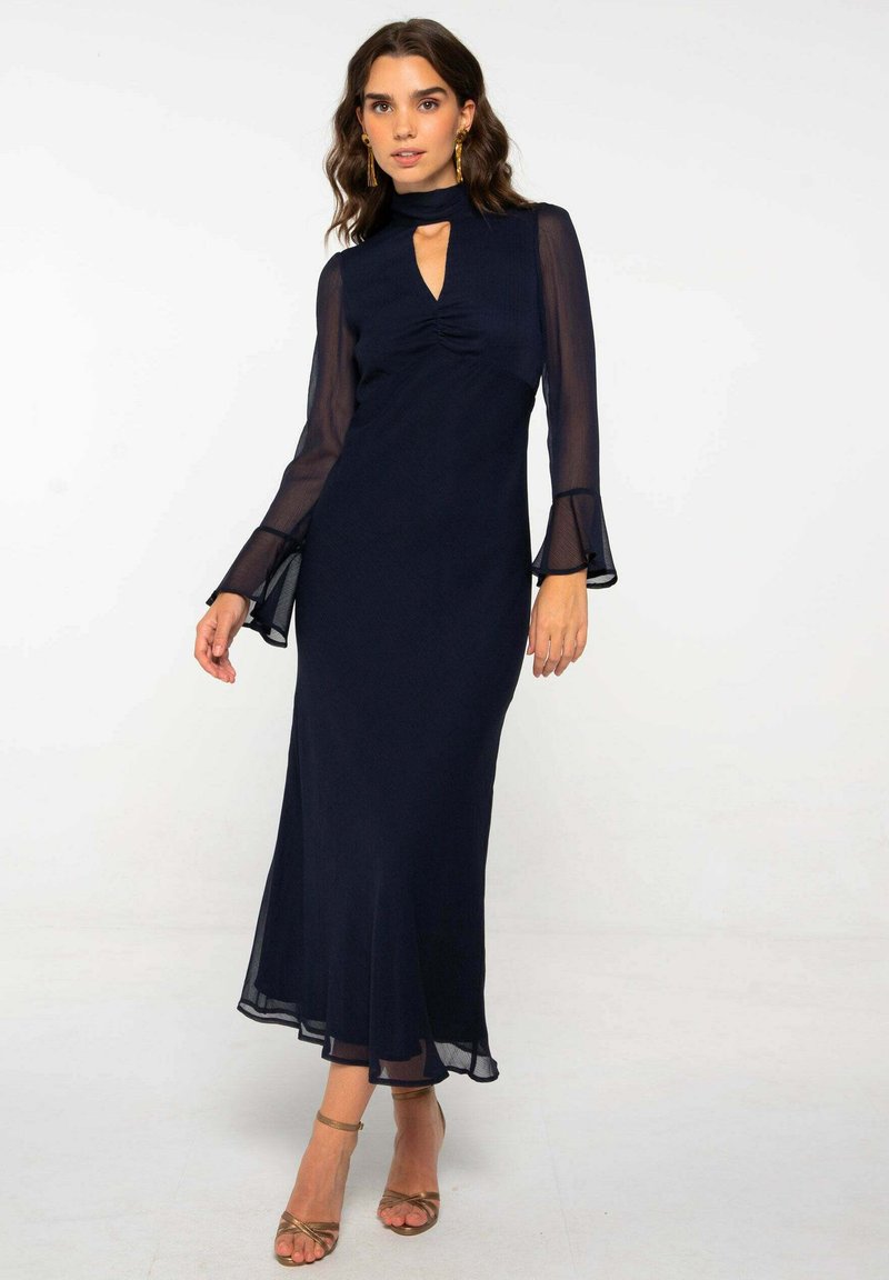 Vestido azul marino con mangas largas transparentes, cuello alto y un escote en forma de llave. Con un dobladillo acampanado y un material texturizado. Combinado con tacones dorados con correas.