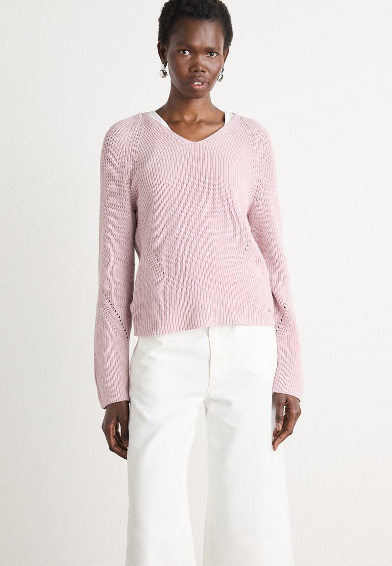 Lichtroze gebreide trui met een V-hals, geribbelde textuur en opengewerkte accenten op de mouwen. In combinatie met witte broek met wijd uitlopende pijpen.