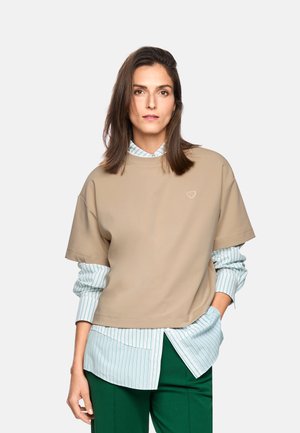 Woman with shoulder-length brown hair wearing a beige short-sleeve top over a white and green striped shirt and green pants, standing with one hand in pocket.