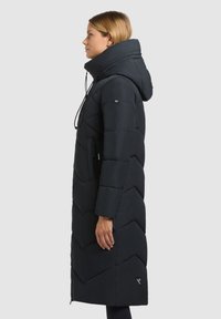 Lange zwarte pufferjas met een hoge kraag en ritssluiting, voorzien van chevronstiksels, een verstelbare trekkoordkap en zijzakken.