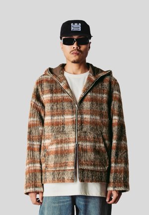 Homme portant une casquette noire, des lunettes de soleil foncées, une veste zippée à carreaux dans des tons marron et beige, une chemise blanche et un jean bleu sur un fond gris uni.