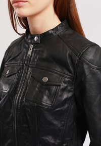 Black leather jacket with a high collar, zip closure, and two chest pockets featuring snap buttons. Smooth texture and tailored fit.