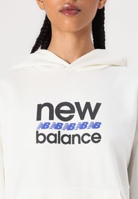 Kremkrāsa kapuce, ar izteiktu melno un zilo logo "new balance" krūšu daļā. Mīksts materiāls, brīvs piegriezums.