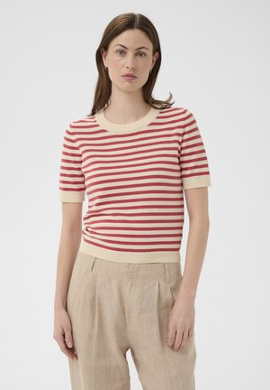 Donna con capelli castani lisci indossa una maglietta a maniche corte a righe rosse e crema e pantaloni beige a vita alta, in piedi davanti a uno sfondo semplice.