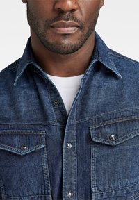 Camisa de mezclilla azul oscuro con collar puntiagudo, dos bolsillos en el pecho y botones de presión plateados, que presenta un tejido texturizado y un diseño ajustado.