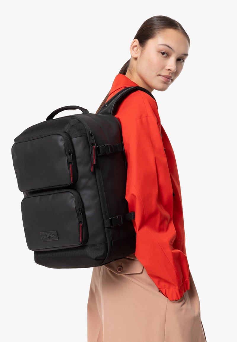 Mochila negra con dos bolsillos frontales, correas de hombro acolchadas y acentos de cremallera roja. Hecha de material duradero y suave, cuenta con un asa superior.