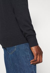 Donkergrijze gebreide trui met geribbelde manchetten, details van een hand die rust op blauwe spijkerbroek, met een casual, aansluitende stijl.