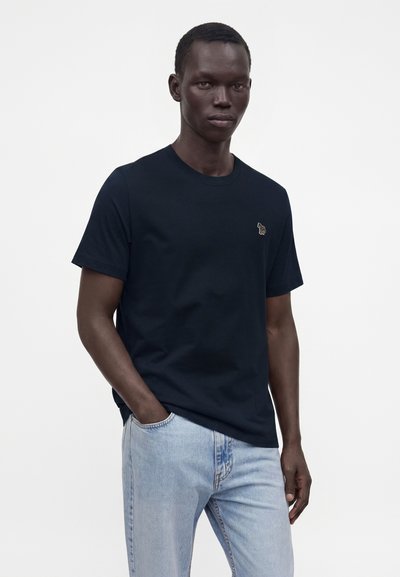 Άνδρας που φοράει απλό μαύρο κοντομάνικο T-shirt με μικρό κεντητό λογότυπο στο στήθος και ανοιχτό μπλε τζιν, στέκεται με το ένα χέρι στην τσέπη.
