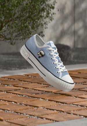 Baskets en toile bleu clair avec lacets blancs et bout renforcé blanc, logo tête de mort noir et blanc, flottant au-dessus d'un sol en carrelage en bois à l'extérieur.