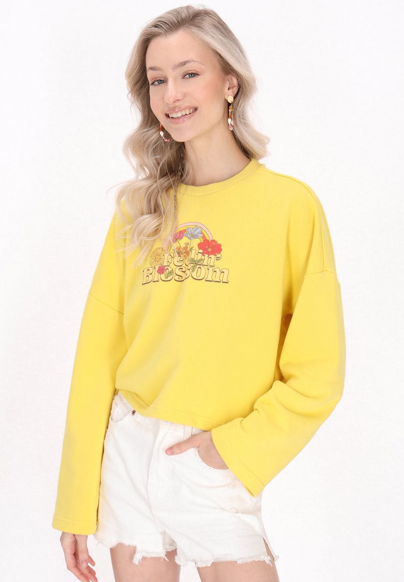 Žlutá oversized mikina s květinovou grafikou a nápisem "Feelin' Blossom", spojená s bílými nařasenými šortkami. Měkká bavlněná tkanina.