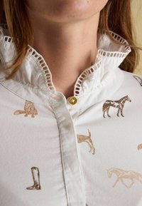 Biela košeľa s naozaj prepracovaným čipkovým lemovaním, s hnedými vzormi zvierat a topánok, a so zlatým gombíkom pri krku.