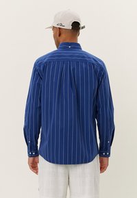 Niebieska koszula z długim rękawem w paski, z kołnierzykiem zapinanym na guziki, plisowanym tyłem i mankietami na guziki. Materiał wydaje się gładki, z tkanym wzorem.