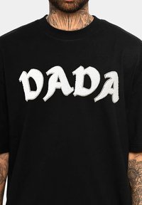 Schwarzes T-Shirt mit weißem, gesticktem Text "DADA" in einer auffälligen, unregelmäßigen Schriftart. Kurze Ärmel, Rundhalsausschnitt und weicher Stoff.