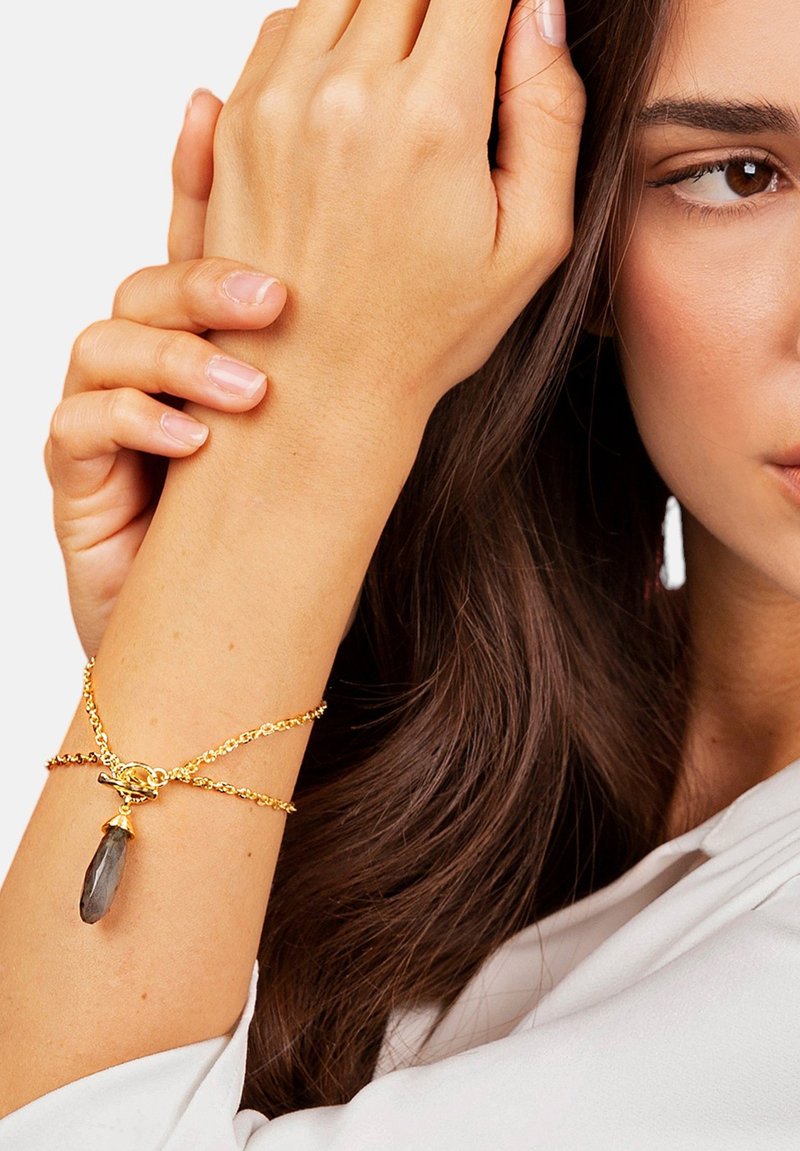 Pulsera de cadena de oro con un prominente colgante de cristal gris, mostrada en una muñeca sobre un fondo neutro.