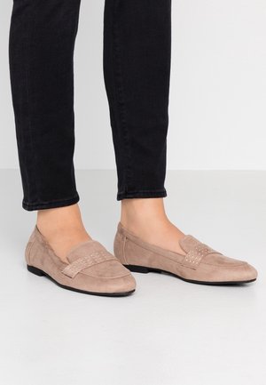 Kvinde iført beige ruskind loafers med små nitter på remmen, kombineret med sorte tætsiddende bukser, står på en lys gulv.