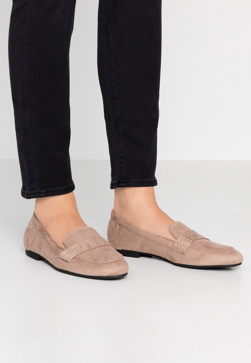 Femme portant des mocassins en daim beige avec de petits clous sur la bride, associés à un pantalon noir ajusté, debout sur un sol clair.