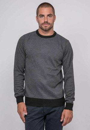Homme aux cheveux courts et barbe, portant un pull à manches longues sombre à motif et un pantalon foncé, debout devant un fond gris uni.
