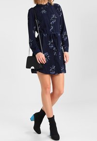 Abito floreale navy con maniche lunghe, colletto e vita con coulisse. Abbinato a stivaletti neri con tacco blu e una piccola borsa nera.