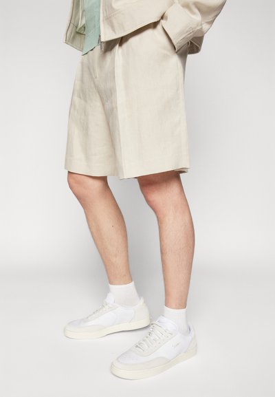 Shorts en lin beige avec des plis, associés à des baskets blanches sportives et des chaussettes blanches mi-mollet. Coupe décontractée, texture lisse et teinte claire.