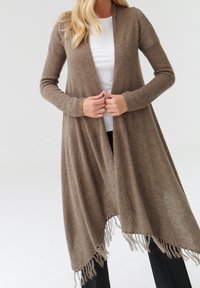 Cardigan largo y abierto de punto en marrón claro, con un patrón texturizado y dobladillo con flecos. Combinado con una blusa blanca y pantalones negros.