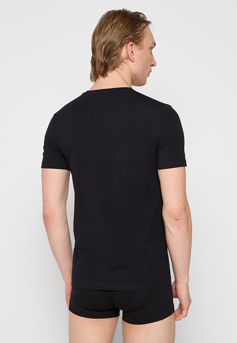 Homme portant un t-shirt noir ajusté à manches courtes et un caleçon noir, se tenant de dos contre un fond clair.