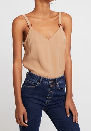 Femme portant un haut beige sans manches à col en V avec des bretelles réglables et un jean taille haute bleu foncé à boutons, debout devant un fond uni.
