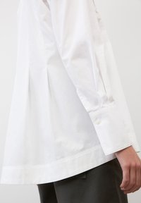 Camisa blanca de manga larga con un ajuste relajado, puños abotonados y un dobladillo ligeramente curvado. Hecha de una tela de algodón suave.