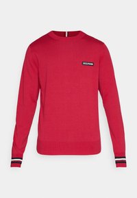 Pull rouge en maille, avec une encolure ronde, des manches longues et un patch logo sur la poitrine. Les poignets sont ornés d'accents rayés.