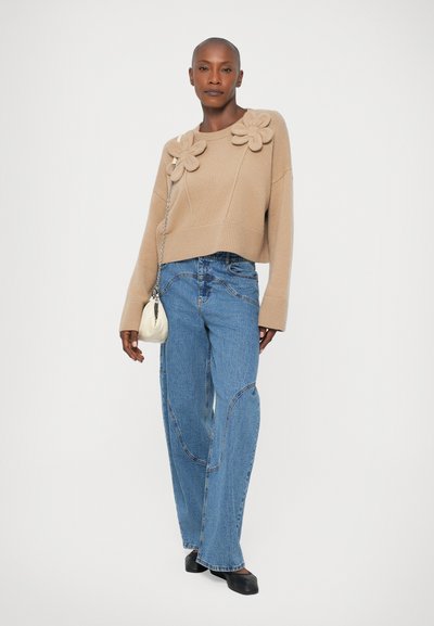 Beige stickad tröja med tre tygblommor på vänster axel, ihop med ljusblå vida denimjeans och en liten vit handväska.