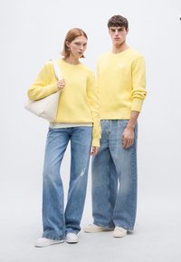 Κίτρινα πλεκτά πουλόβερ με ριμπ μανσέτες, σε συνδυασμό με ανοιχτό μπλε τζιν ψηλόμεσα. Η γυναίκα κρατάει μια λευκή τσάντα. Και οι δύο φορούν λευκά αθλητικά παπούτσια.