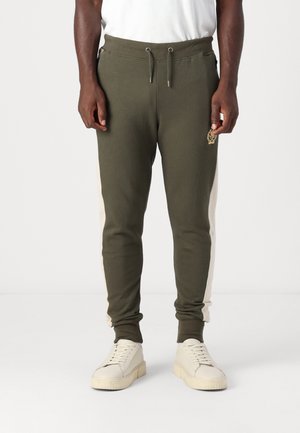 Homme portant un pantalon de jogging vert olive avec des bandes blanches sur les côtés et des baskets beiges, debout devant un fond clair uni.