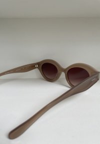 Occhiali da sole in plastica marrone con montatura arrotondata e lenti scure. Presentano asticelle sottili con un logo discreto. Texture liscia, design moderno.