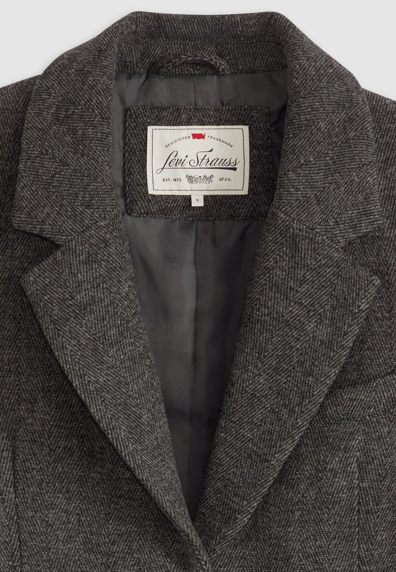 Grå fiskbensmönstrad kappa med klädda slag, med en slät innerfoder. Etiketten visar "Levi Strauss" med storleksmarkering.