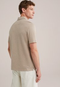 Camisa polo beige de manga corta con un diseño de punto texturizado, un collar clásico y aberturas laterales en el dobladillo, combinada con pantalones de color claro.