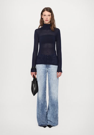 Tricou turtleneck din plasă navy, cu un model texturat, asortat cu blugi evazați, de culoare albastru deschis, ușor deteriorați. Modelul ține o geantă mică, neagră.