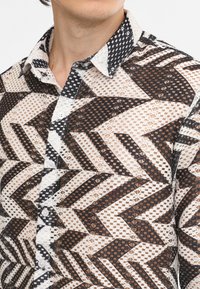 Camicia a maniche corte con motivo geometrico in nero, bianco e marrone. Caratterizzata da tessuto testurizzato, colletto e chiusura con bottoni davanti.