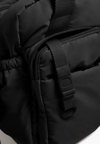 Črn oblazinjen nahrbtnik s gladko teksturo, z žepi na zadrgo in nastavljivimi naramnicami s plastično sponko.