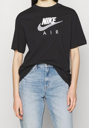 Osoba ubrana w czarną koszulkę Nike Air wpuszczoną do jasnoniebieskich, wysokich dżinsów, z ramionami luźno opuszczonymi wzdłuż ciała.