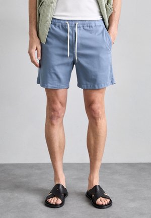 Personne portant un short bleu clair à cordon, un t-shirt blanc, une chemise vert clair et des sandales noires à bout ouvert, debout sur un sol gris.