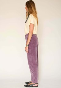 Un pantalon en velours côtelé violet avec une coupe droite, associé à un t-shirt jaune clair. Des ballerines noires, pointues, avec des détails en œillets.