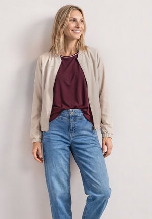 Blonde vrouw met een beige jas over een bordeaux shirt en blauwe jeans, staat en glimlacht terwijl ze opzij kijkt tegen een effen muur.