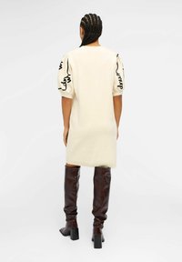 Vestido color crema con mangas abullonadas que presenta bordados abstractos en negro. Modelo vistiendo botas de charol marrón hasta la rodilla con un tacón grueso.