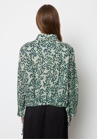 Kobieta z długimi, brązowymi włosami, ubrana w jasnoskalny bluzkę w ciemnozielony wzór w kropki, widziana z tyłu, oraz czarną spódnicę.