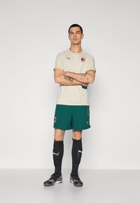 Beżowa koszulka sportowa z logo, zielone krótkie spodenki, czarne skarpetki do kolan oraz czarne buty sportowe. Prosty design z delikatnymi akcentami i detalami.