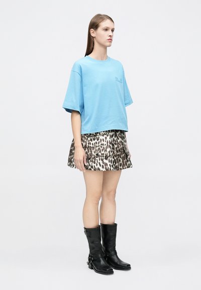 Νεαρή γυναίκα που φοράει μπλε φαρδιά μπλούζα, μίνι φούστα με λεοπάρ σχέδιο και τσέπες cargo, καθώς και μαύρες δερμάτινες μπότες μέχρι τη μέση της γάμπας, πάνω σε λευκό φόντο.