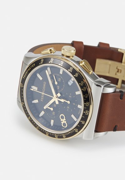 Orologio cronografo con quadrante blu navy, dettagli in oro, cassa in acciaio inossidabile argentato e cinturino in pelle marrone. Presenta tachimetro e sottodial.