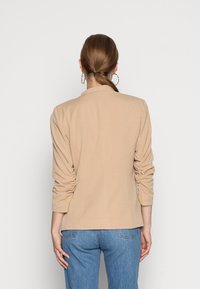 Beige skräddarsydd blazer med jämn textur, med rynkade ärmar och en något strukturerad design. Matchas med ljusblå jeans.