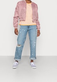 Pink satin bomber jacket, orange speckled top, distressed blue jeans with paint splatters, and purple sneakers. Casual, modern outfit.