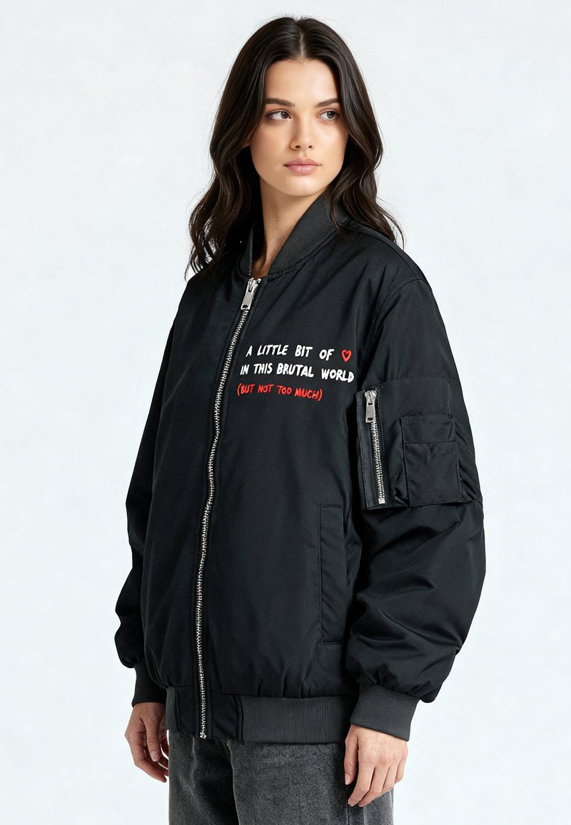 Zwarte bomberjack met een ritssluiting aan de voorkant, geribbelde boorden en zoom. Heeft rood-witte borduursels op de borst en een ritssluiting aan het armzakje.